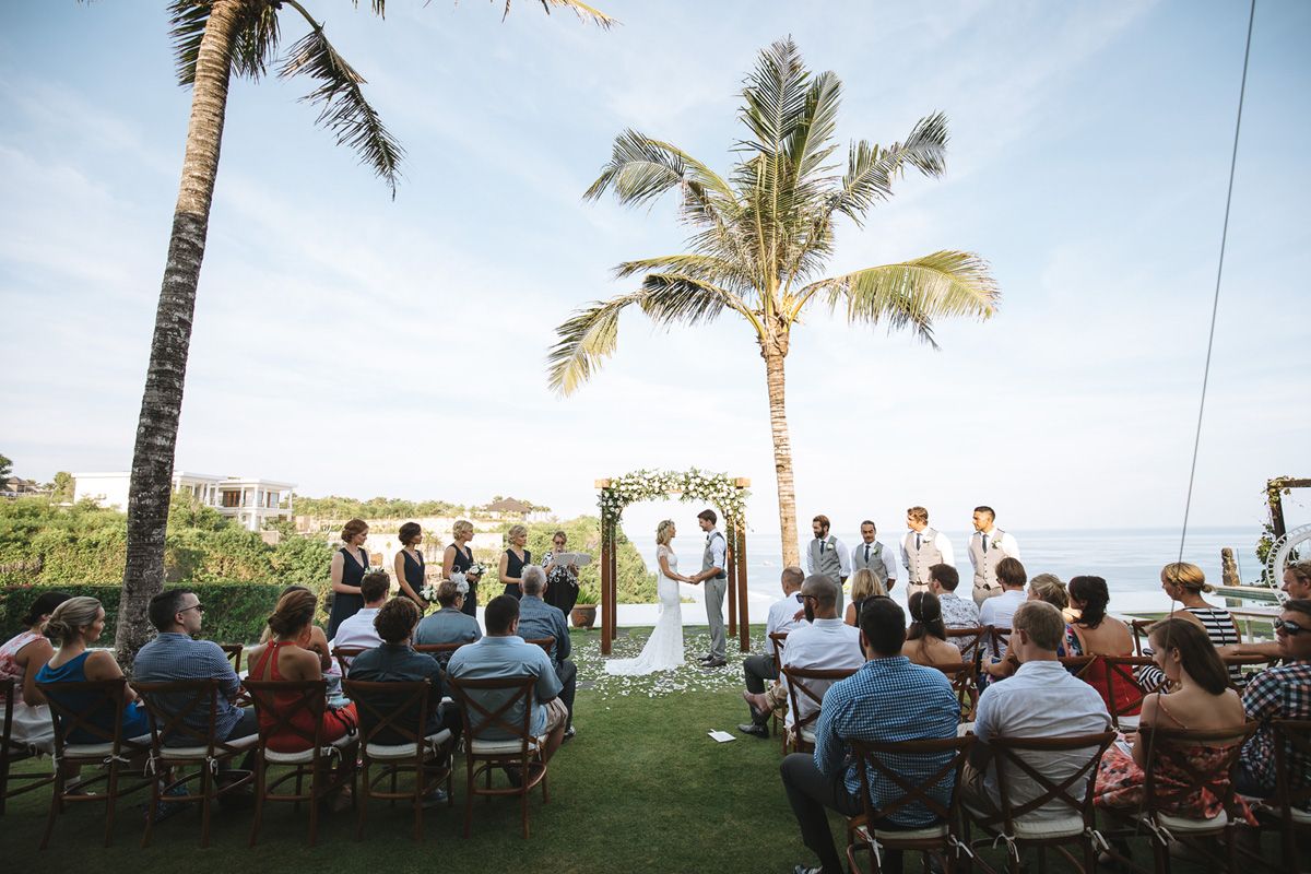 An outdoor wedding ceremony on a scenic oceanfront lawn with palm trees and guests seated in white chairs under a blue sky.