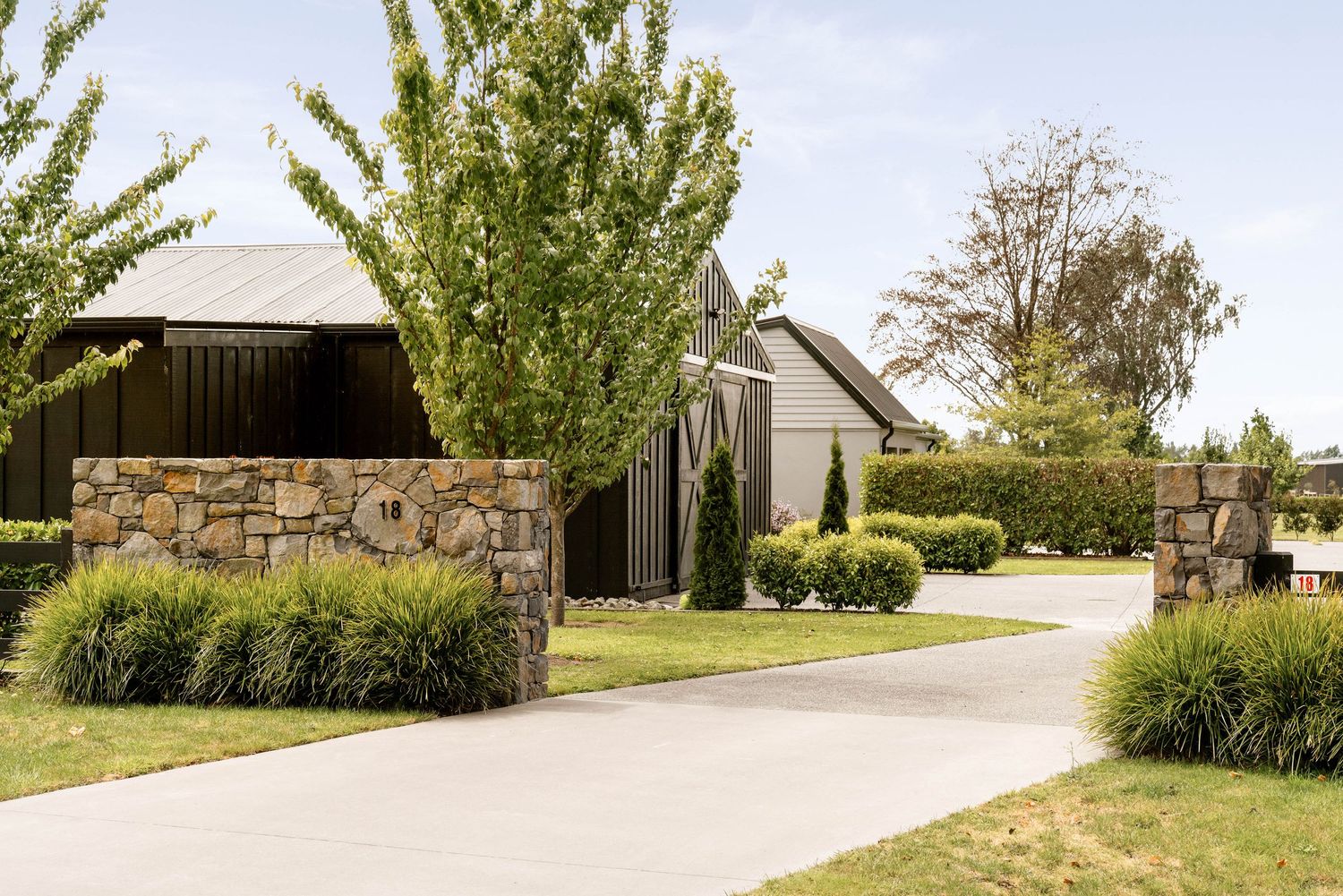Modern farmhouse entrance with stone wall feature and winding driveway surrounded by landscaped gardens.