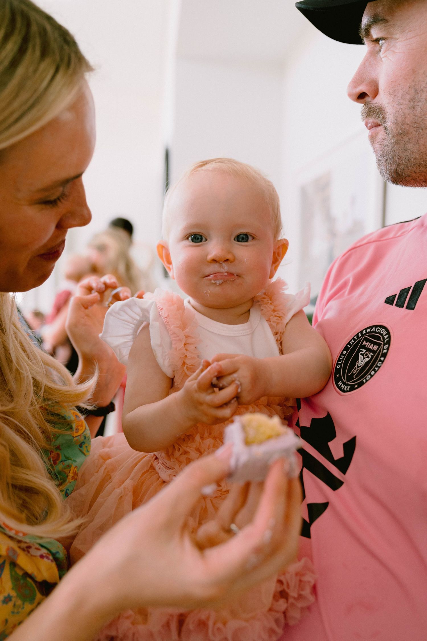 A baby wearing a pink tulle dress smiles while being held during a gathering.