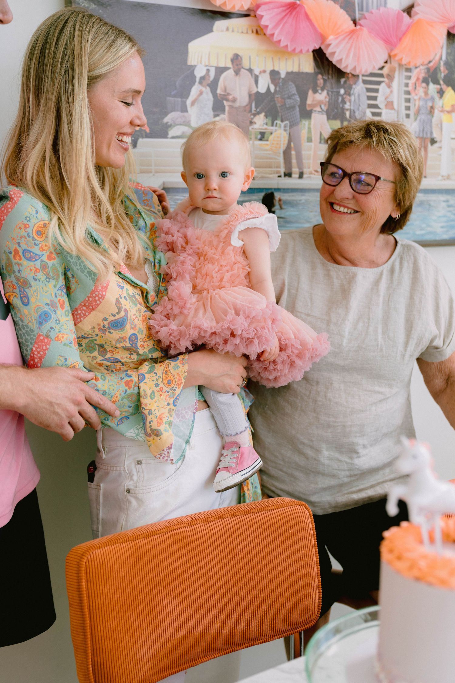 Three generations share a joyful moment at a party decorated with pink tissue paper pom-poms.