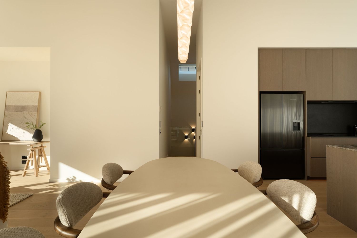 Modern dining room with oval table, beige chairs, black refrigerator, and elegant pendant light in a minimalist interior.