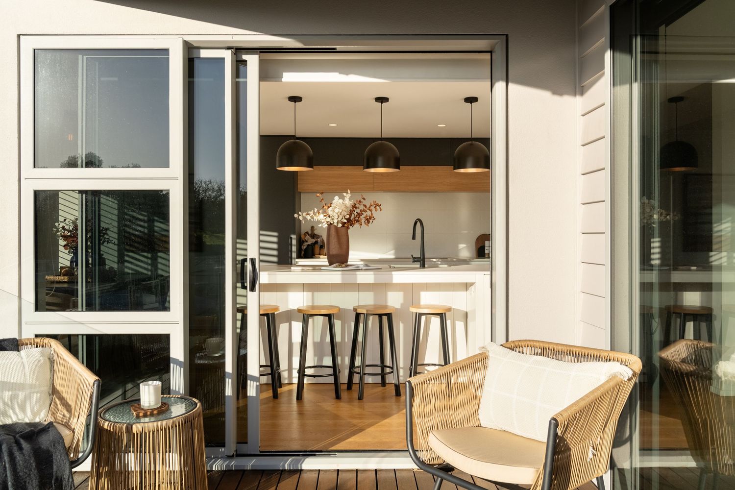 Outdoor patio with wicker chairs opens to modern kitchen featuring bar stools, pendant lights, and white cabinetry.