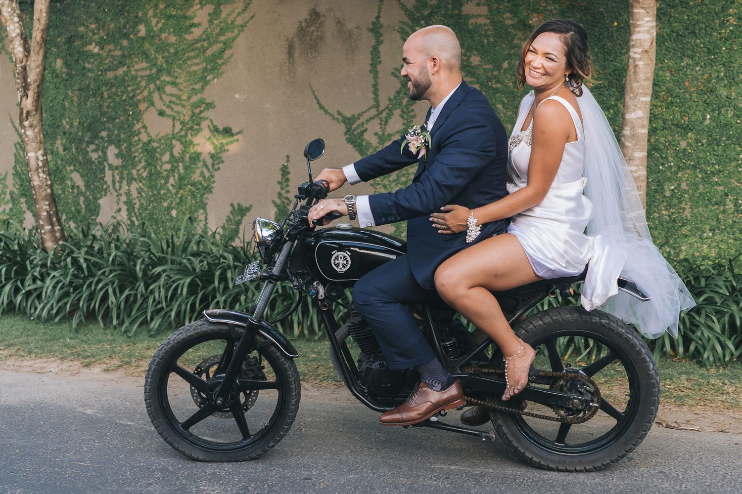 A ride on a black motorcycle shows a couple dressed in wedding attire smiling during their special day.