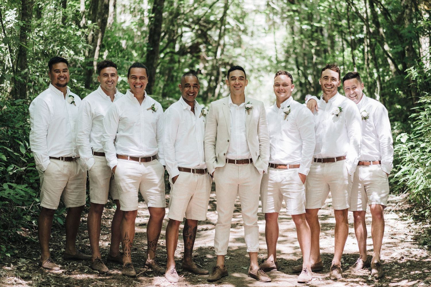 Groomsmen dressed in crisp white shirts and khaki shorts pose together on a forest path.