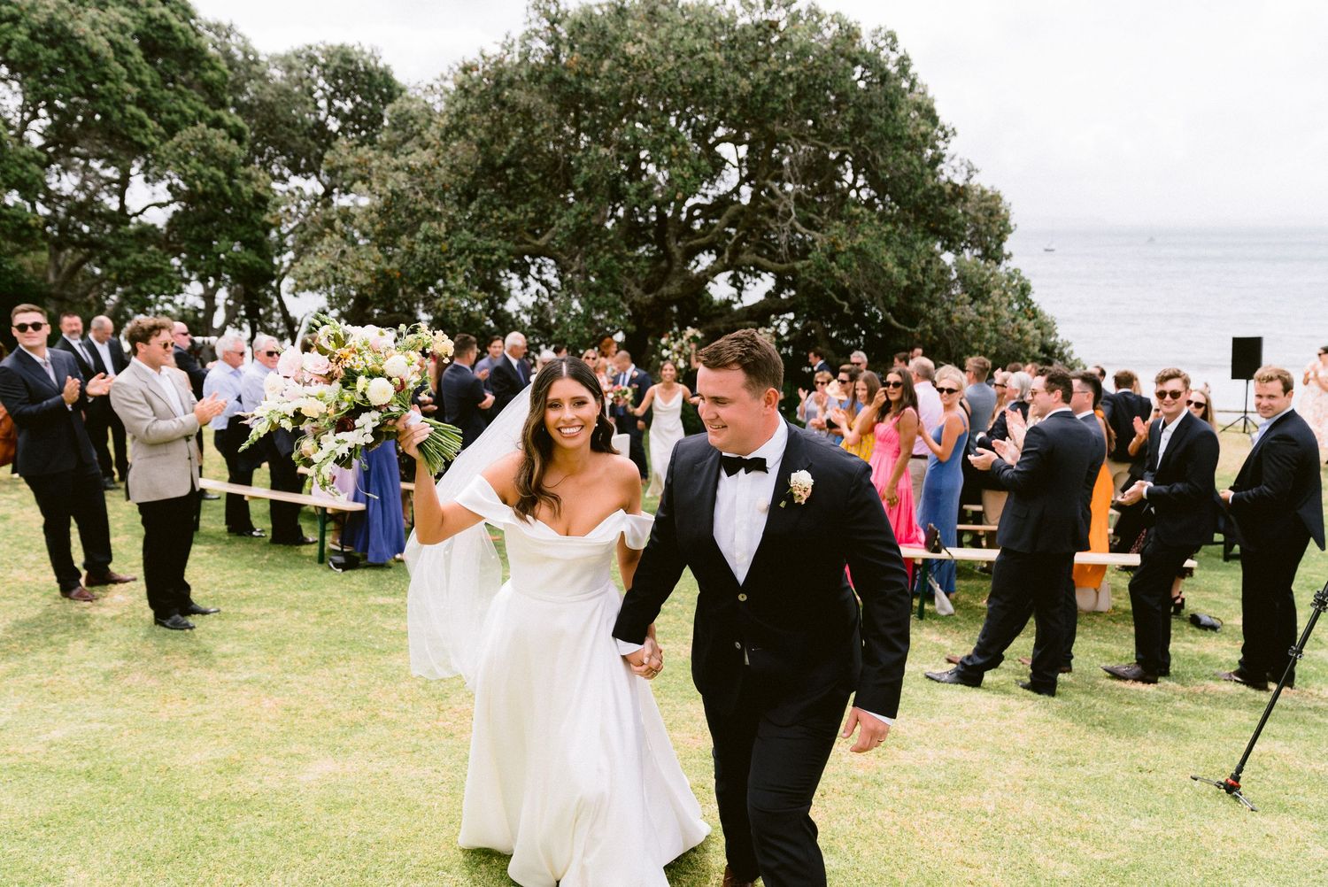 Happy newlyweds exit their wedding ceremony on a grassy lawn while guests look on.