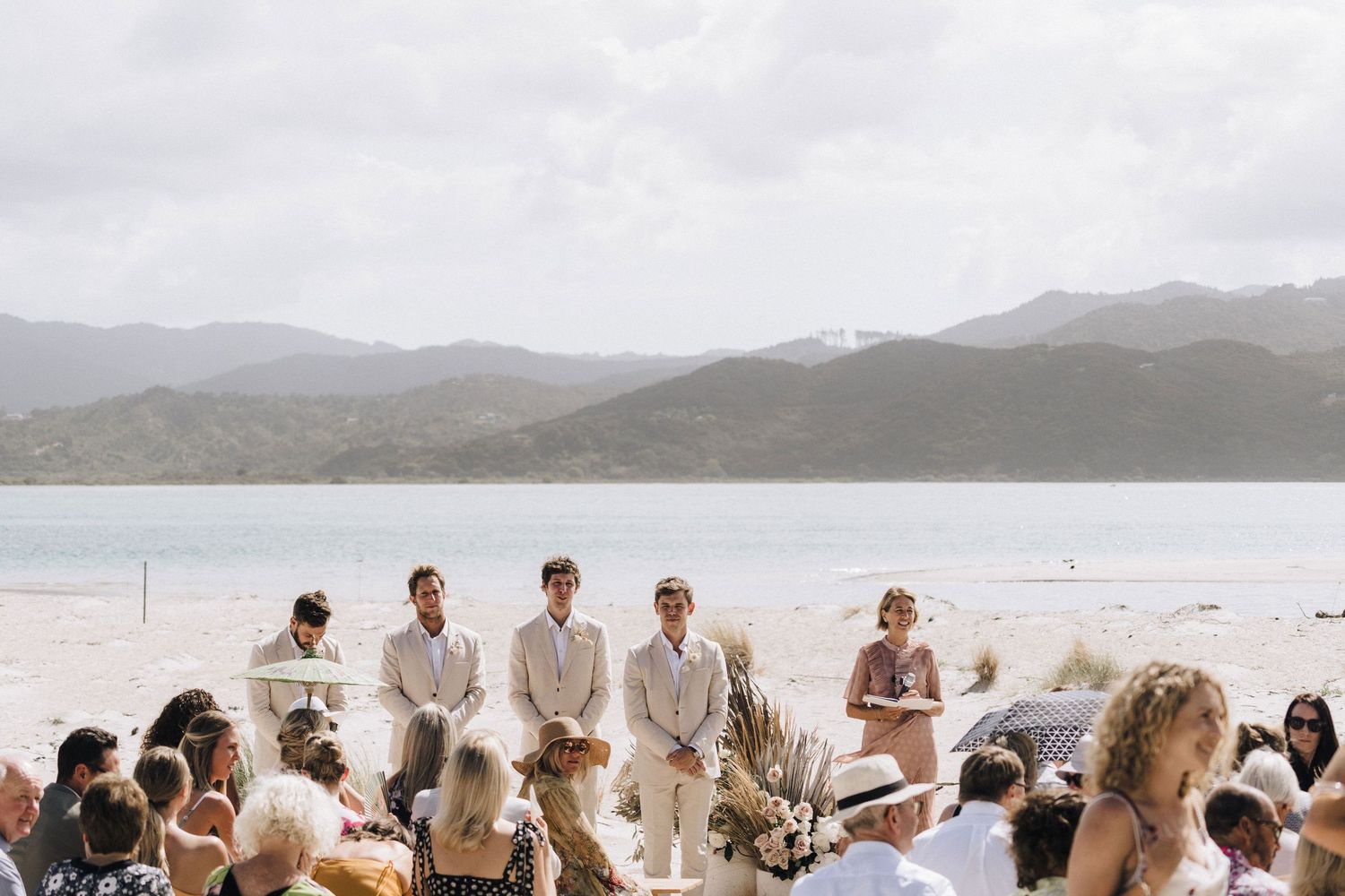 Wedding guests in white attire gather on a scenic beach with mountains in the background.