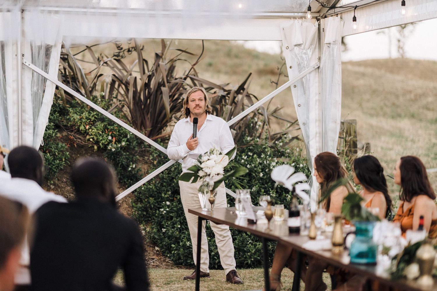 Speaker gives wedding speech under white tent during outdoor reception.