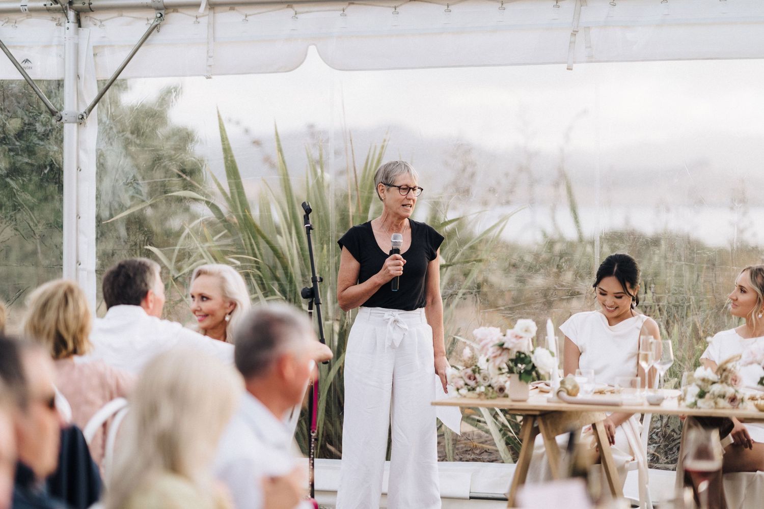 A wedding reception outdoors with people gathered at tables under a tent while someone gives a speech on a sunny day.