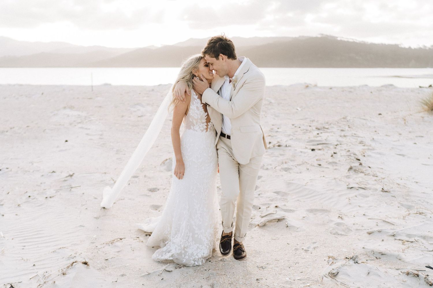 Romantic newlywed couple embracing on a sandy beach with mountains in the background.