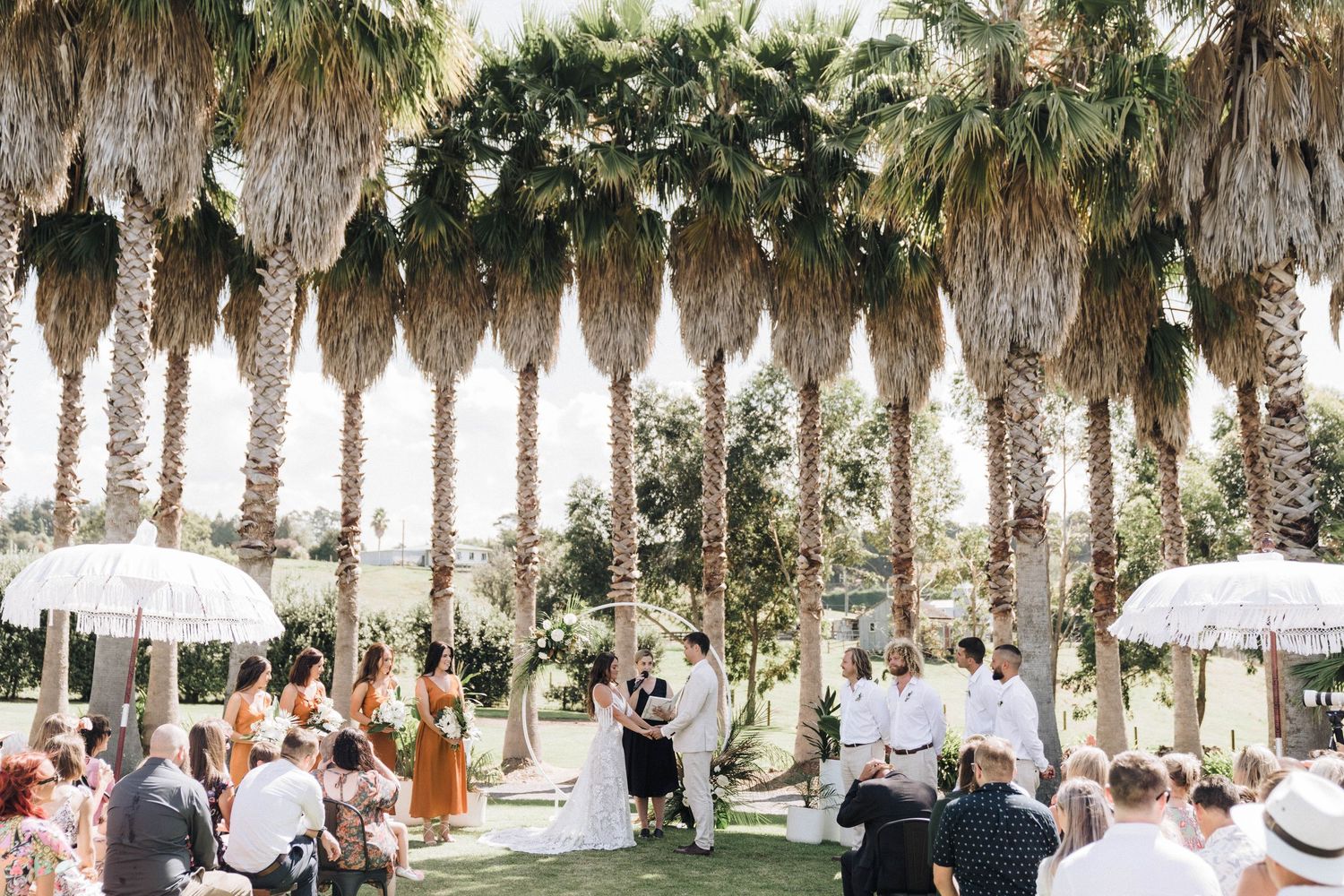 Outdoor wedding ceremony setup with white chairs and umbrellas under towering palm trees.