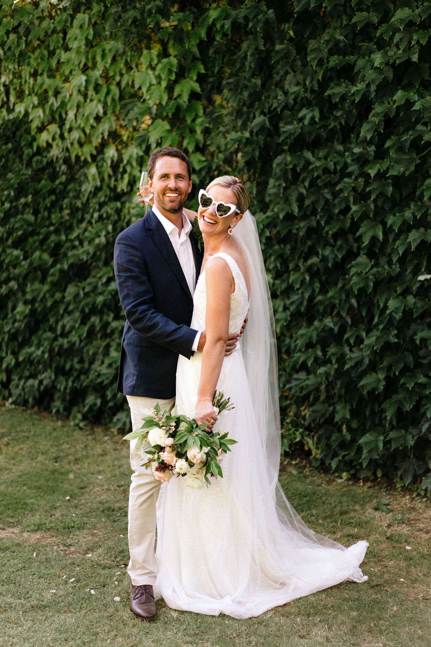 A wedding couple poses against a lush green hedge wall, with the bride wearing sunglasses and holding a floral bouquet.