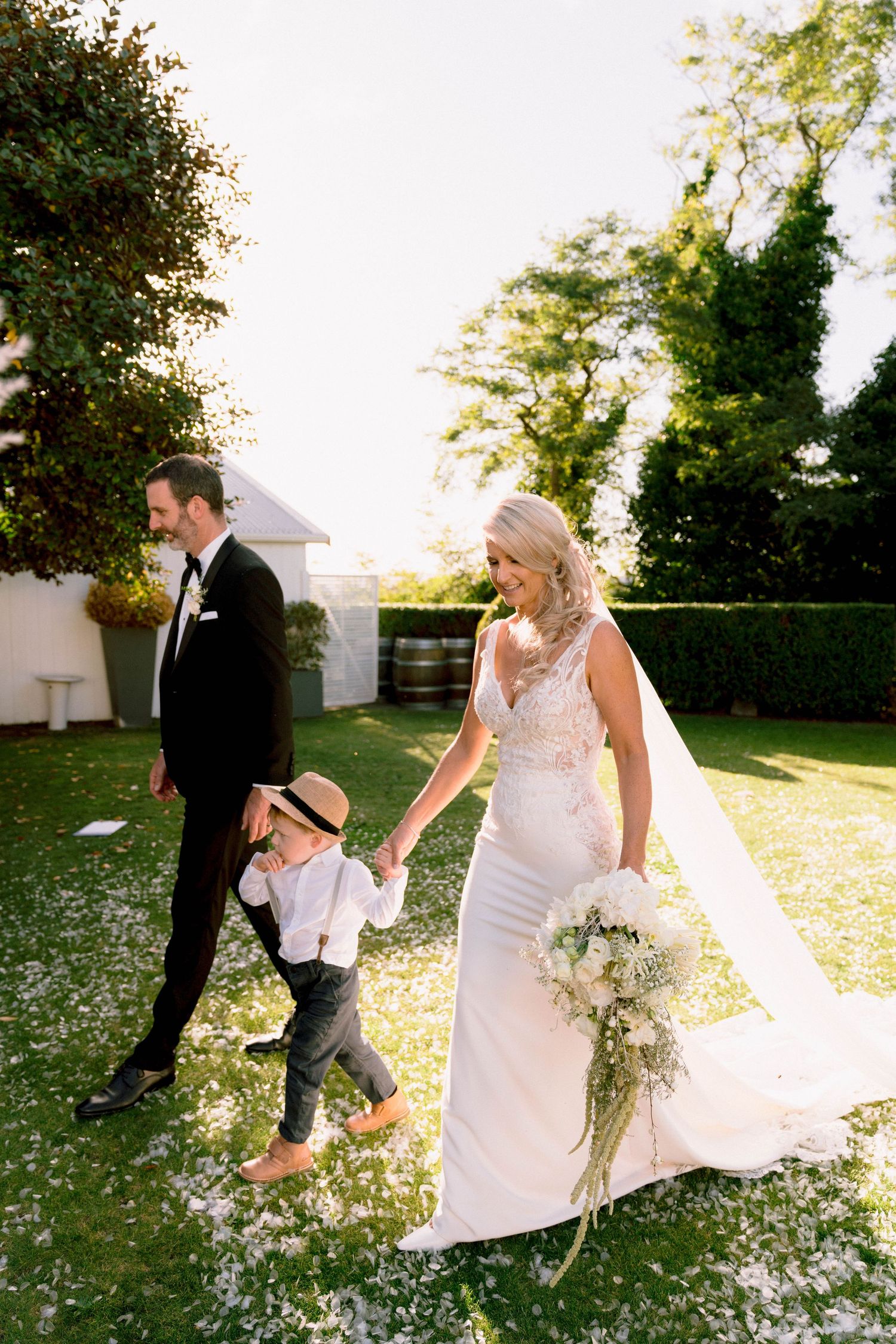 A wedding party walks through scattered flower petals on grass with a young ring bearer in suspenders and a hat.