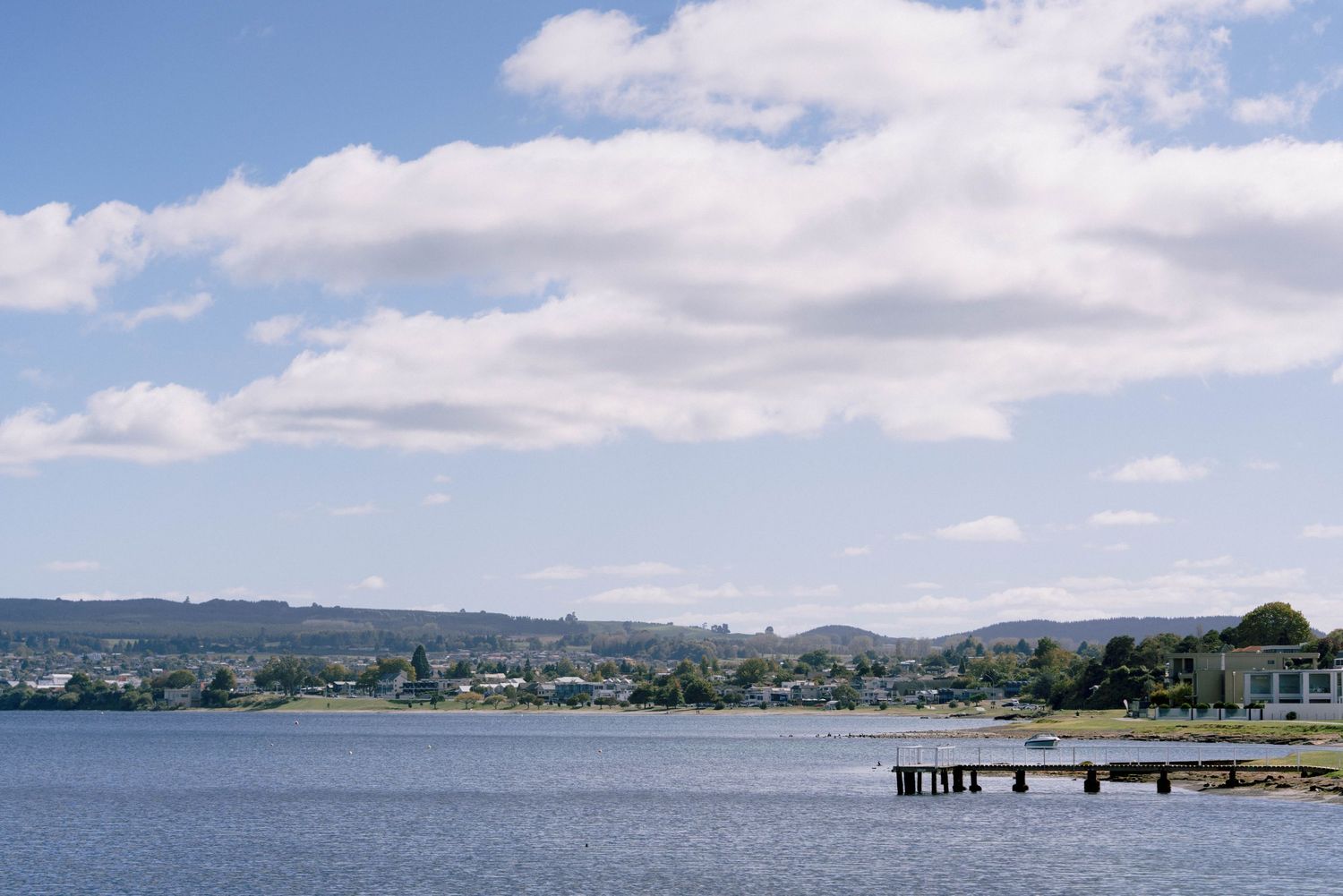 A scenic panoramic view of a coastal town with houses along the shoreline and a wooden pier extending into calm blue waters.
