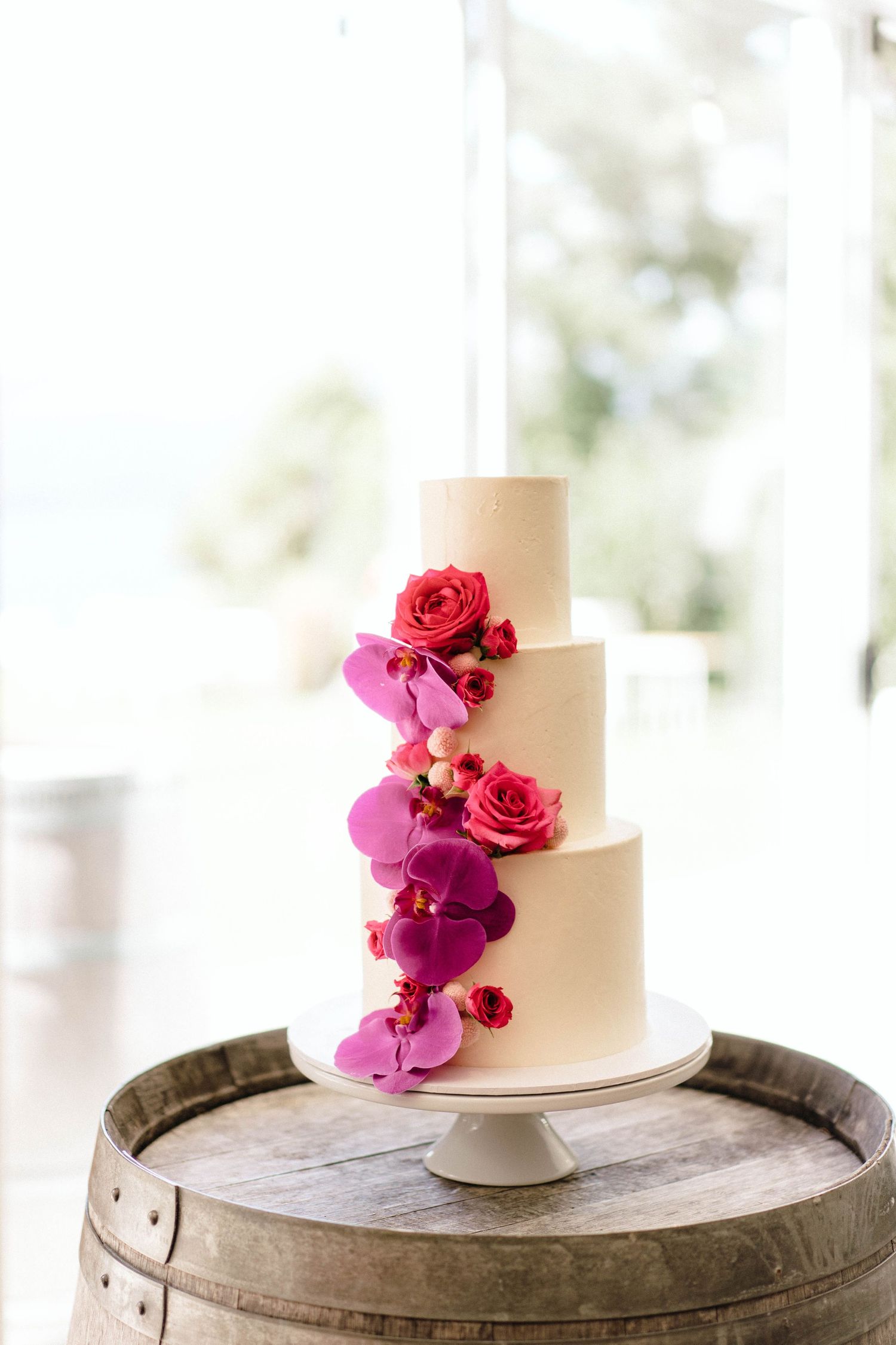 Elegant white wedding cake decorated with vibrant pink and red florals displayed on a rustic wooden barrel.