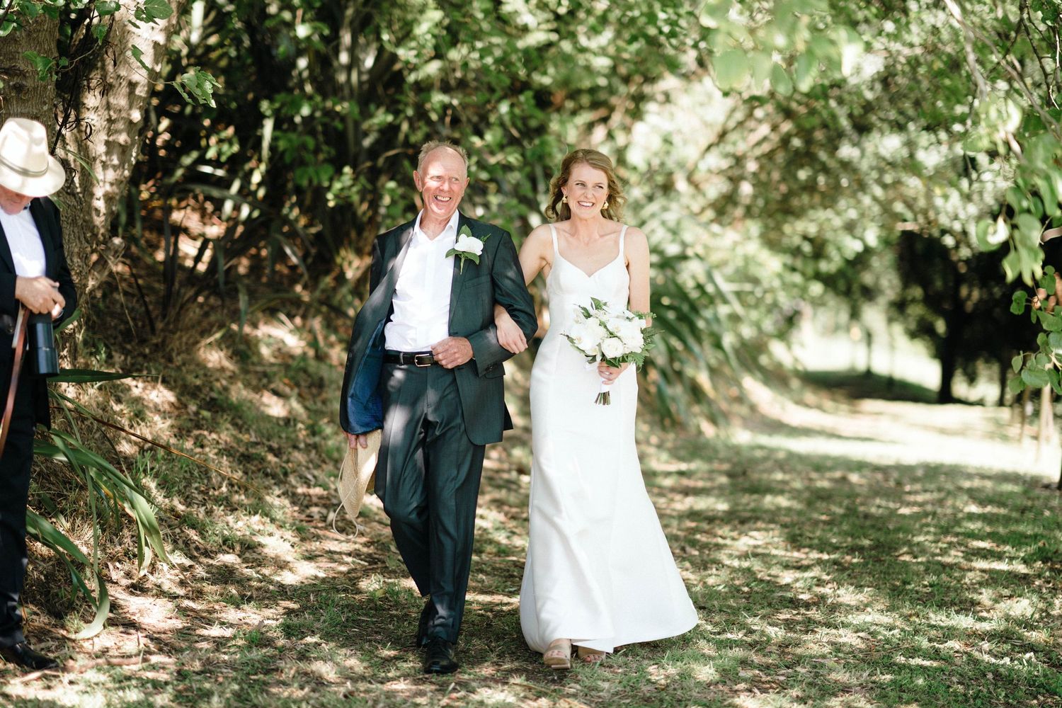 Romantic newlyweds and their loved ones walking through a scenic tree-lined path during a joyful wedding celebration.