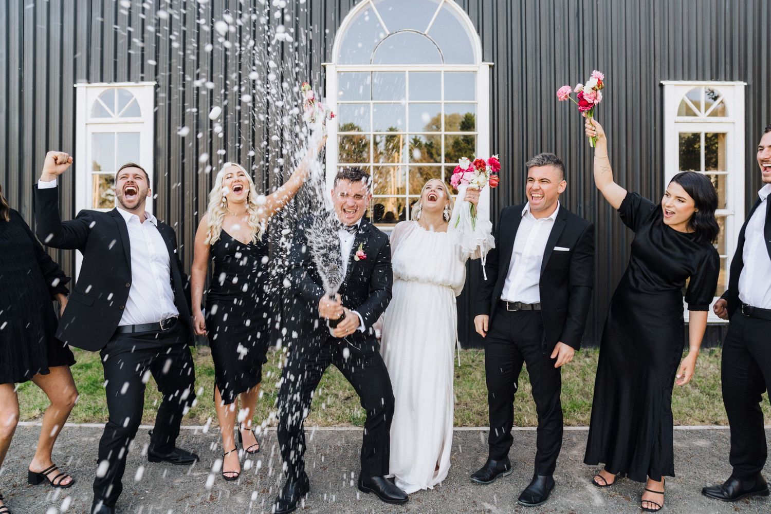 Wedding party celebrates with confetti toss outside venue with black exterior and white windows.