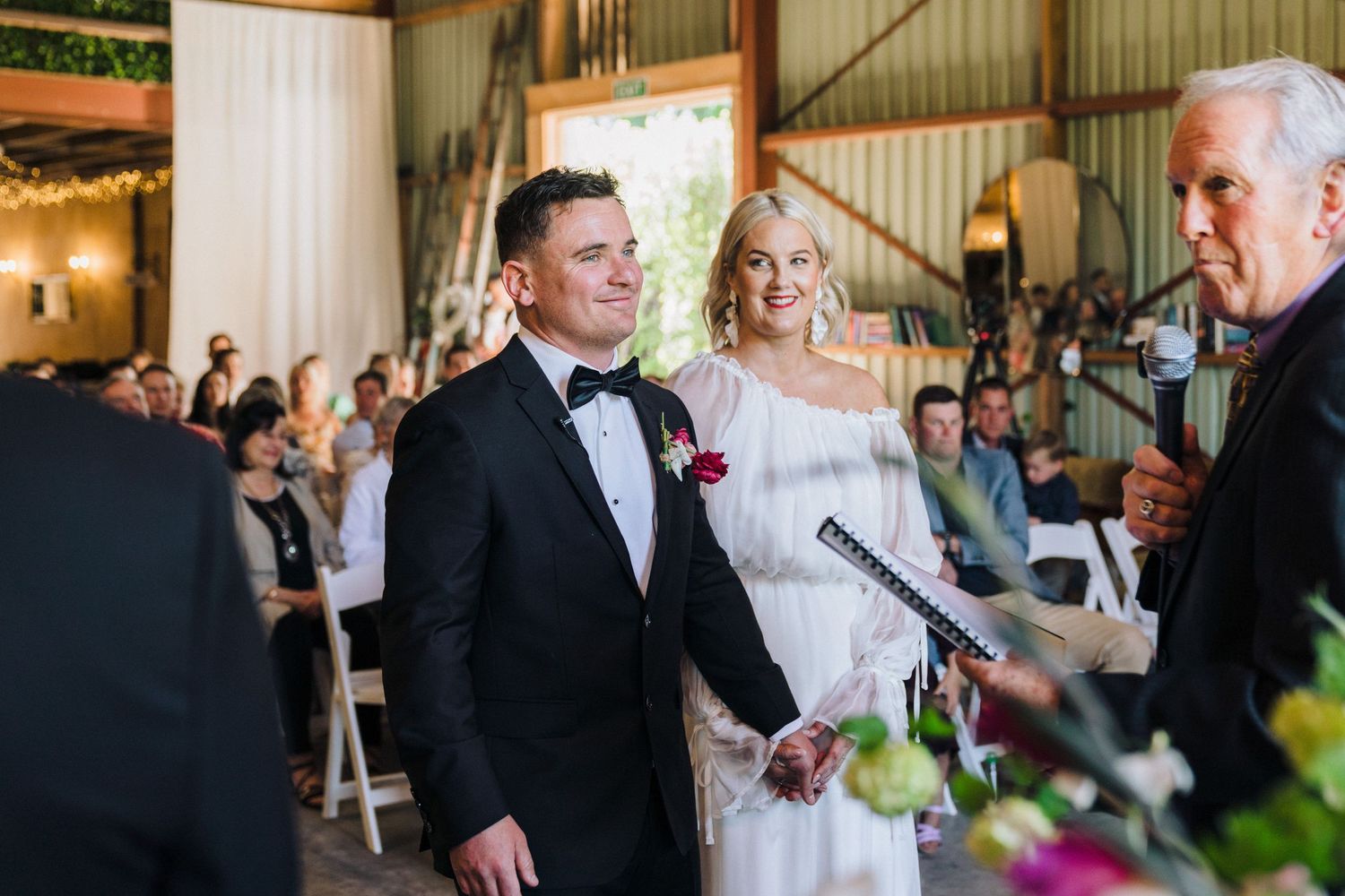 Wedding ceremony in progress inside rustic barn venue with white drapery and floral decorations.