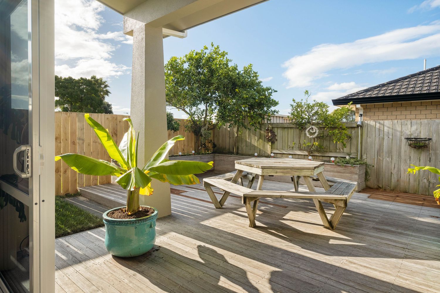 A wooden deck patio with a picnic table and potted banana plant under a covered porch area on a sunny day.
