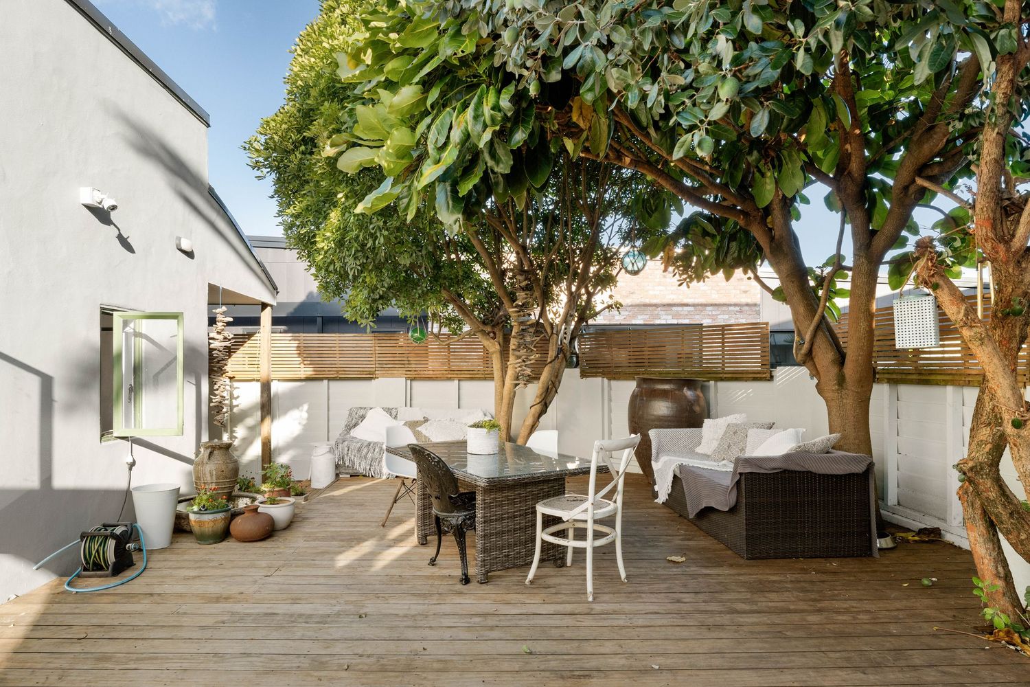 Modern outdoor patio space with wooden deck, comfortable seating, and lush green trees providing shade.