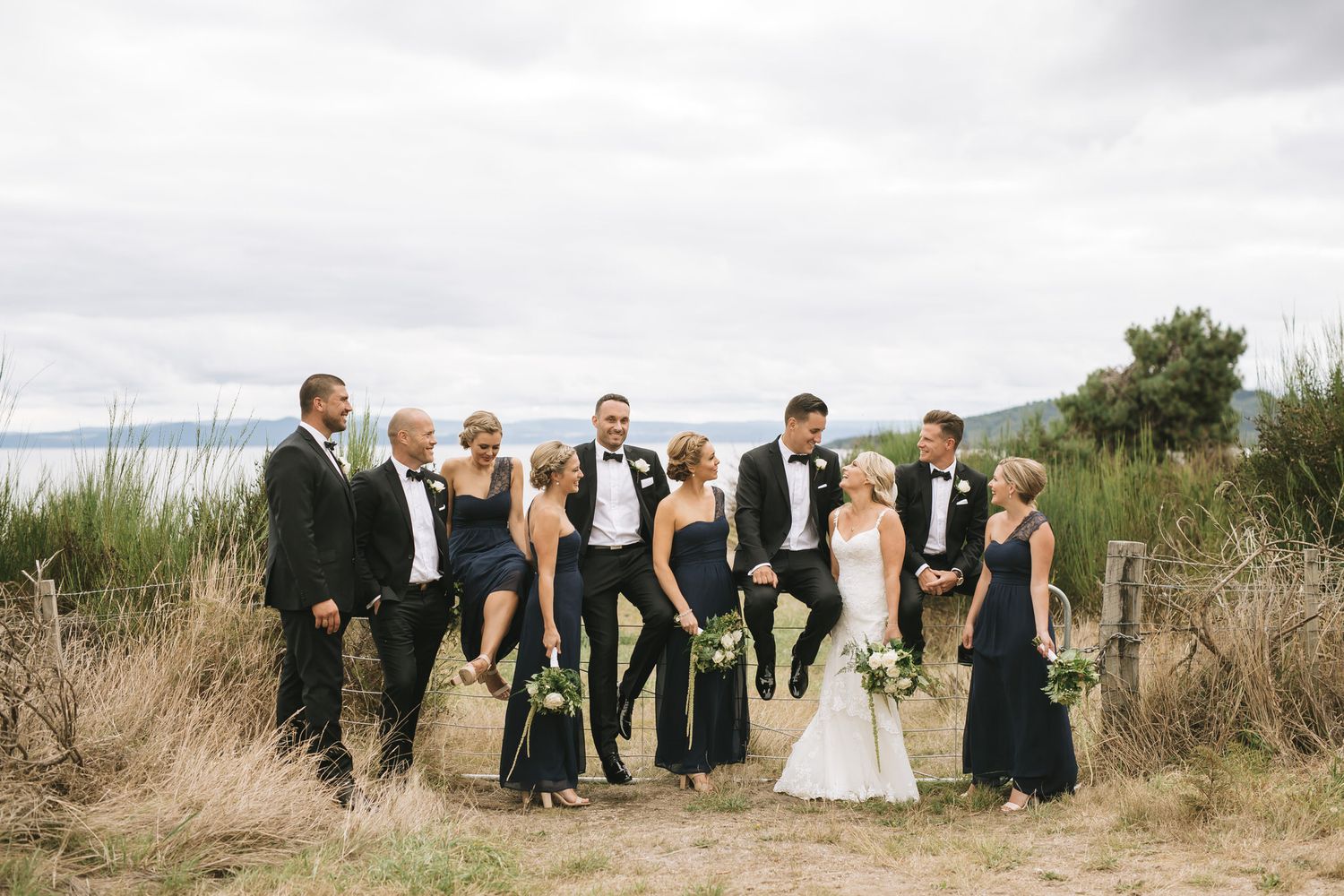 Wedding party in formal attire poses together outdoors on a grassy hill overlooking water and mountains.