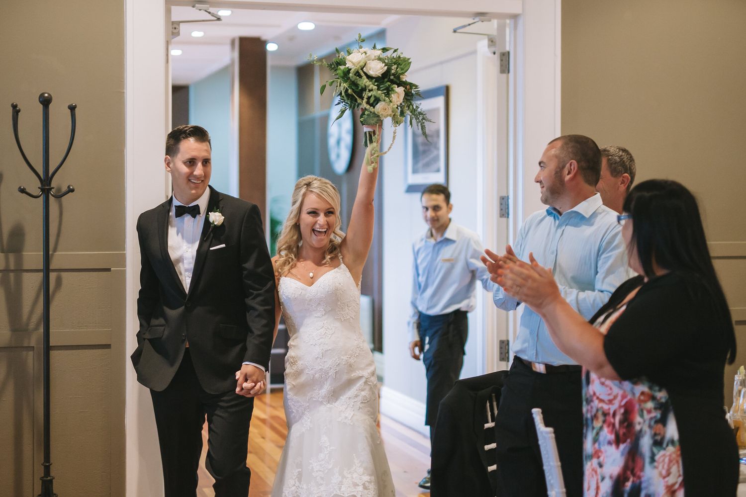 Newlywed couple enters venue hallway while guests applaud and cheer their arrival.