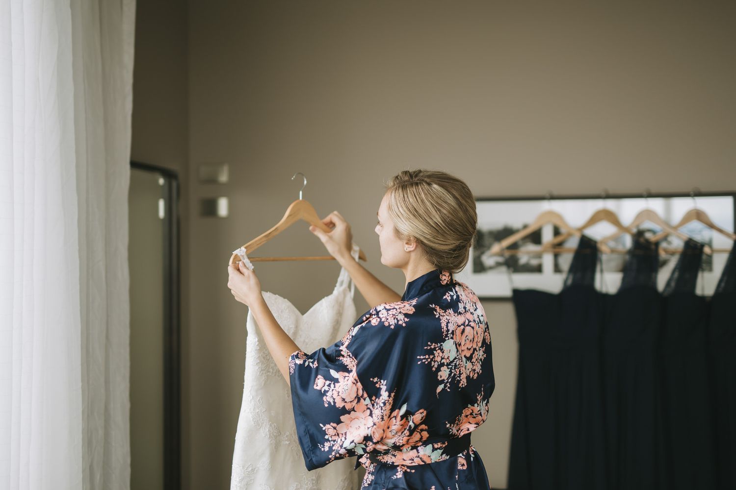 Woman in floral blue robe inspects white dress hanging near clothing rack in bedroom setting.