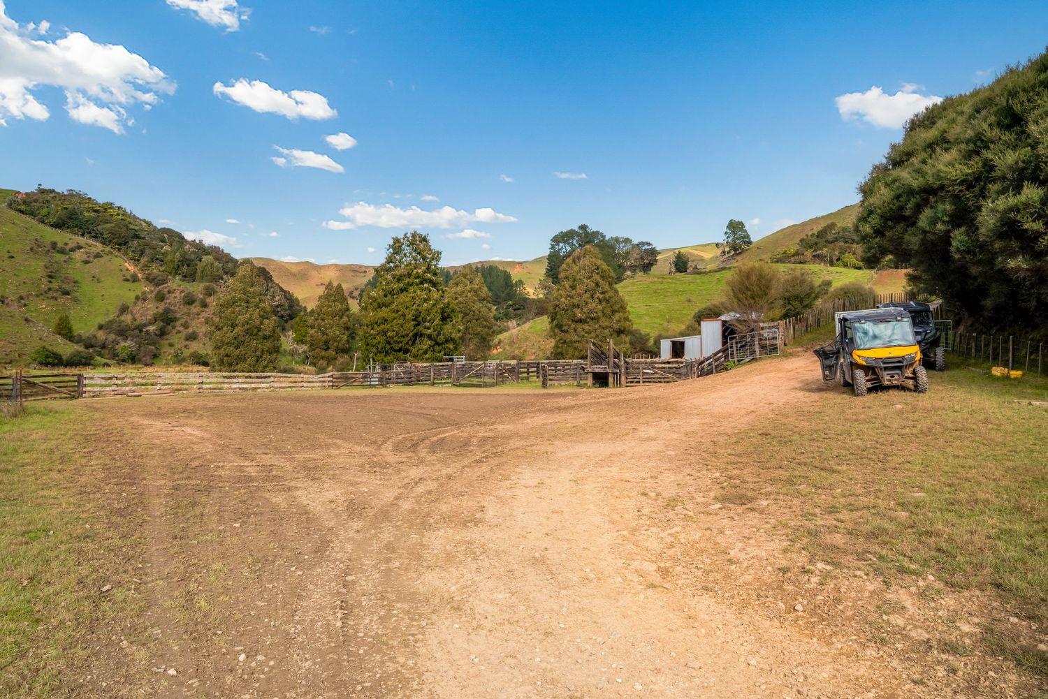 Dirt road intersection in hilly countryside with utility vehicle and farm buildings in distance.