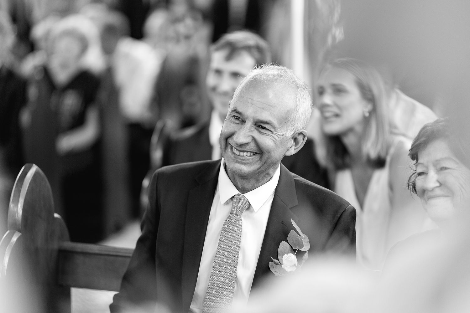 Black and white candid moment captured showing joyful laughter during formal event with guests wearing suits and boutonniere.