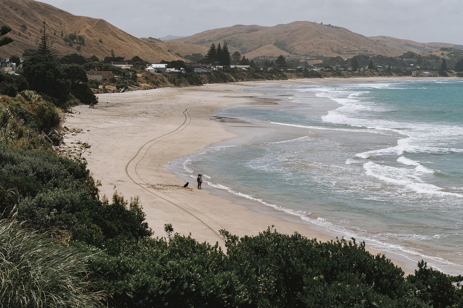 Scenic coastal beach view with rolling hills and turquoise waves lapping at sandy shores on a cloudy day.