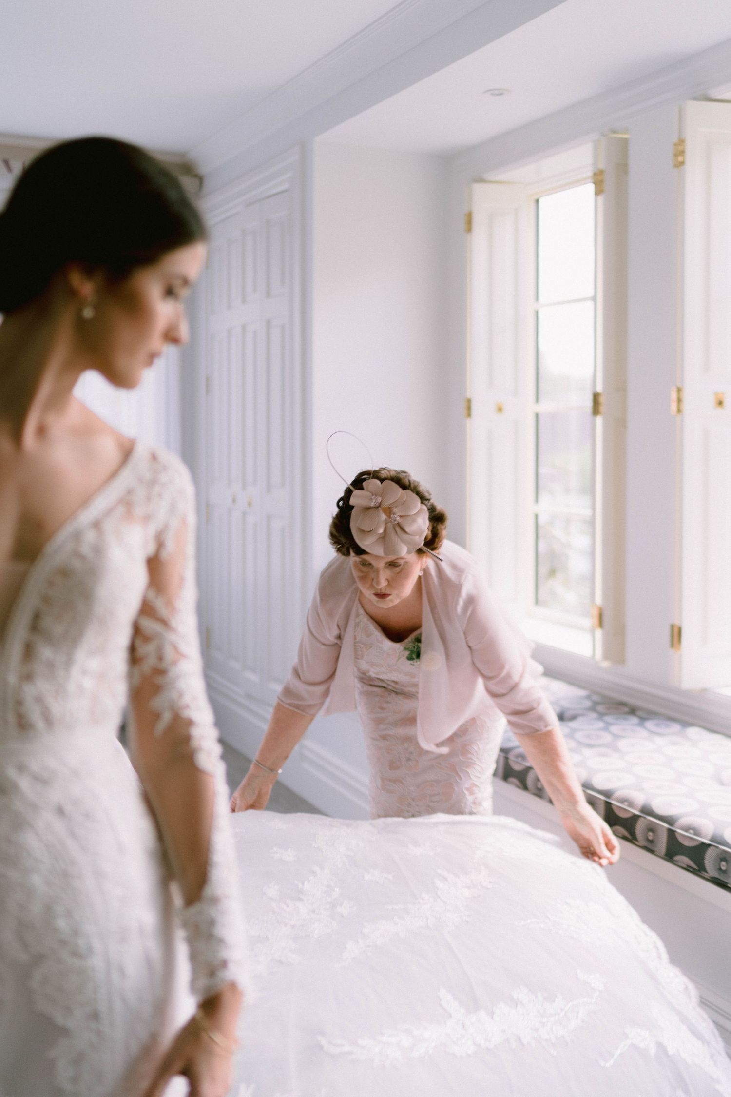 A woman in a white wedding dress being helped with final adjustments in a sunlit dressing room.