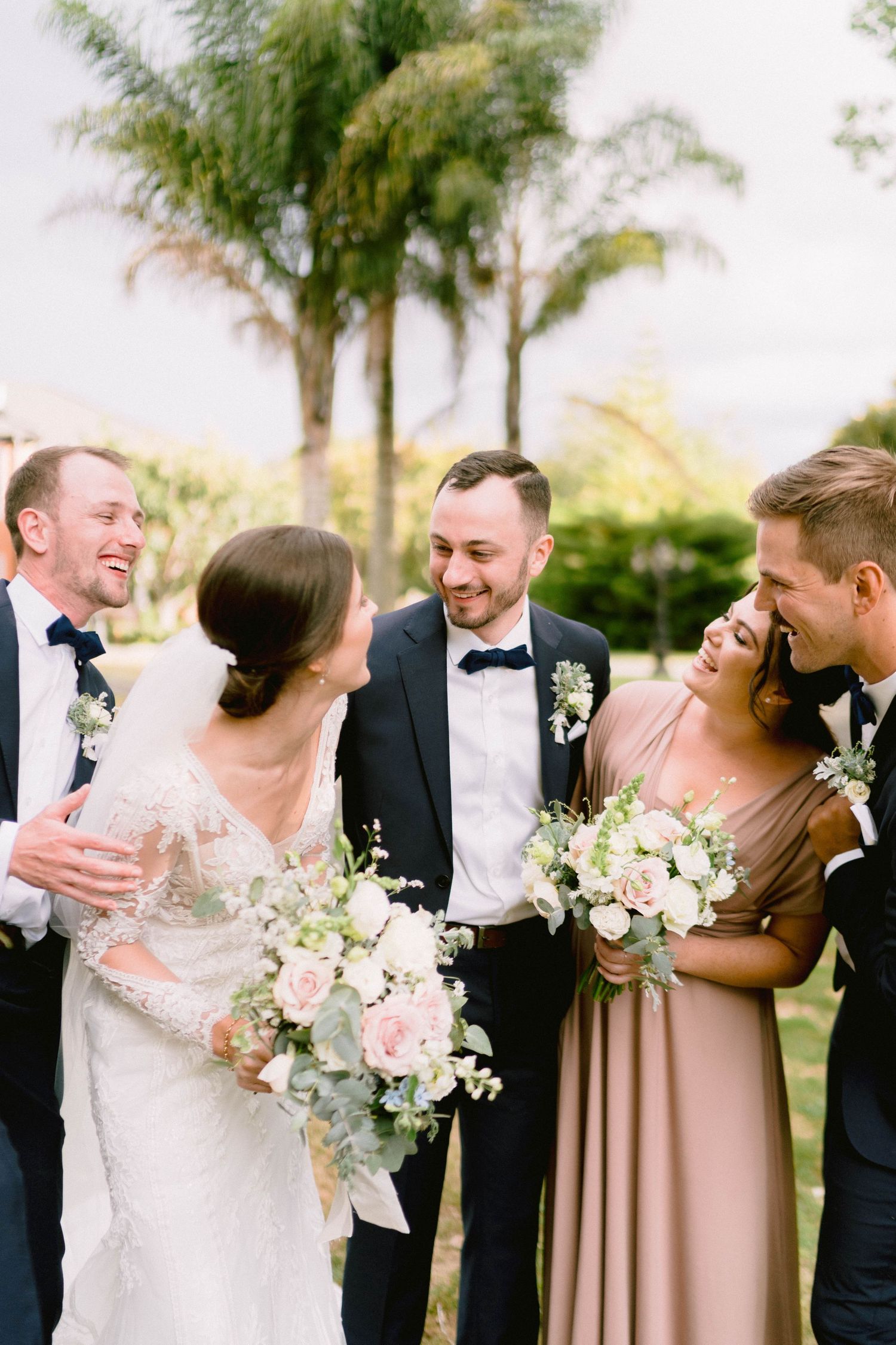 Wedding party laughing together outdoors with white floral bouquets and palm trees in background.