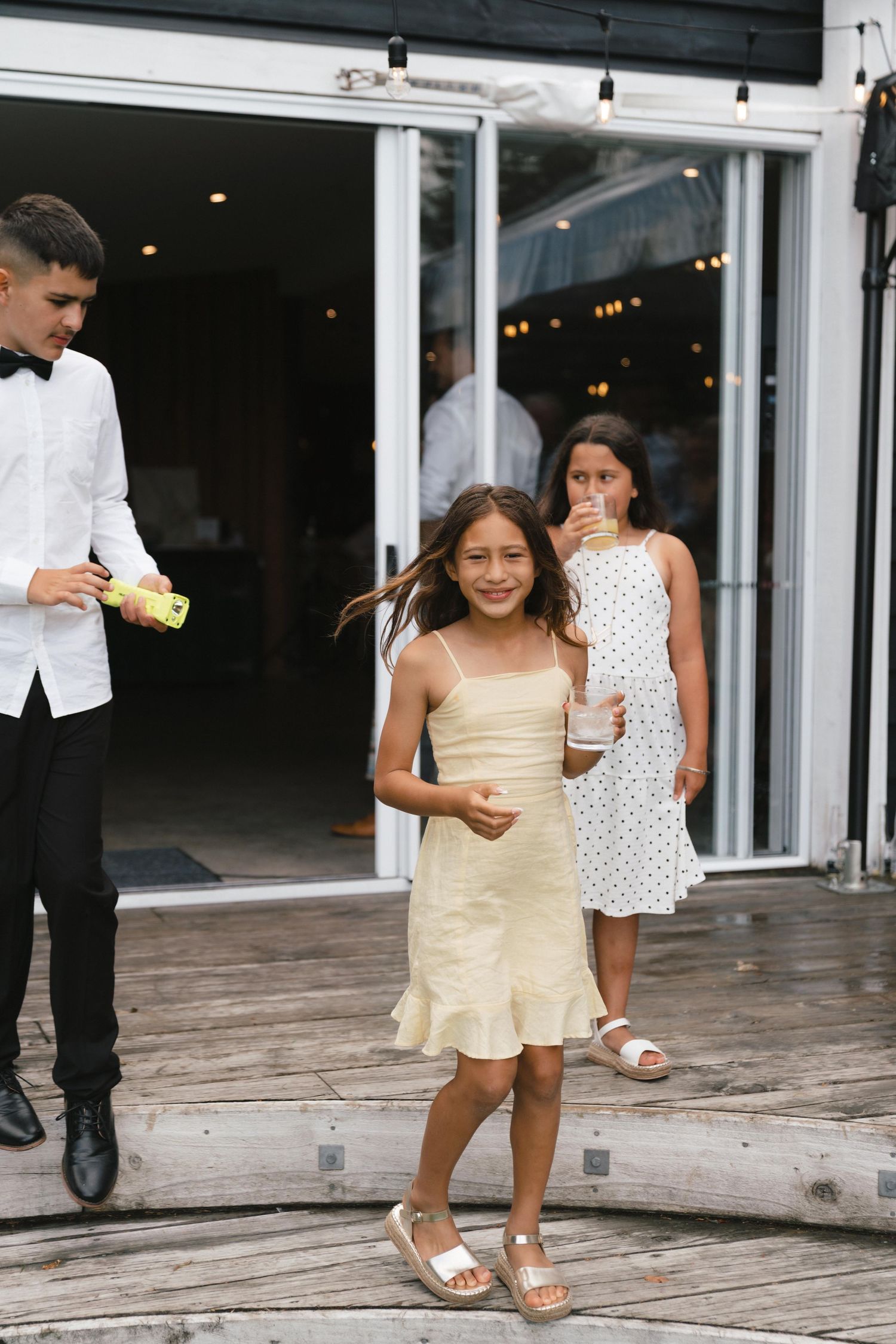 Young wedding guests in casual attire gather on a wooden deck outside sliding glass doors.
