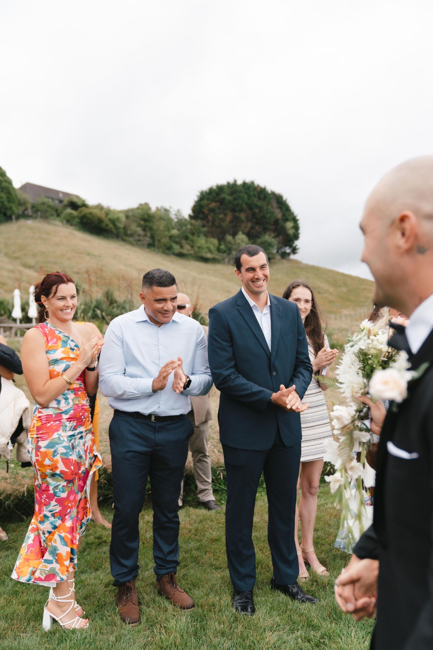 Wedding guests in formal wear gather on green hillside for group photos.