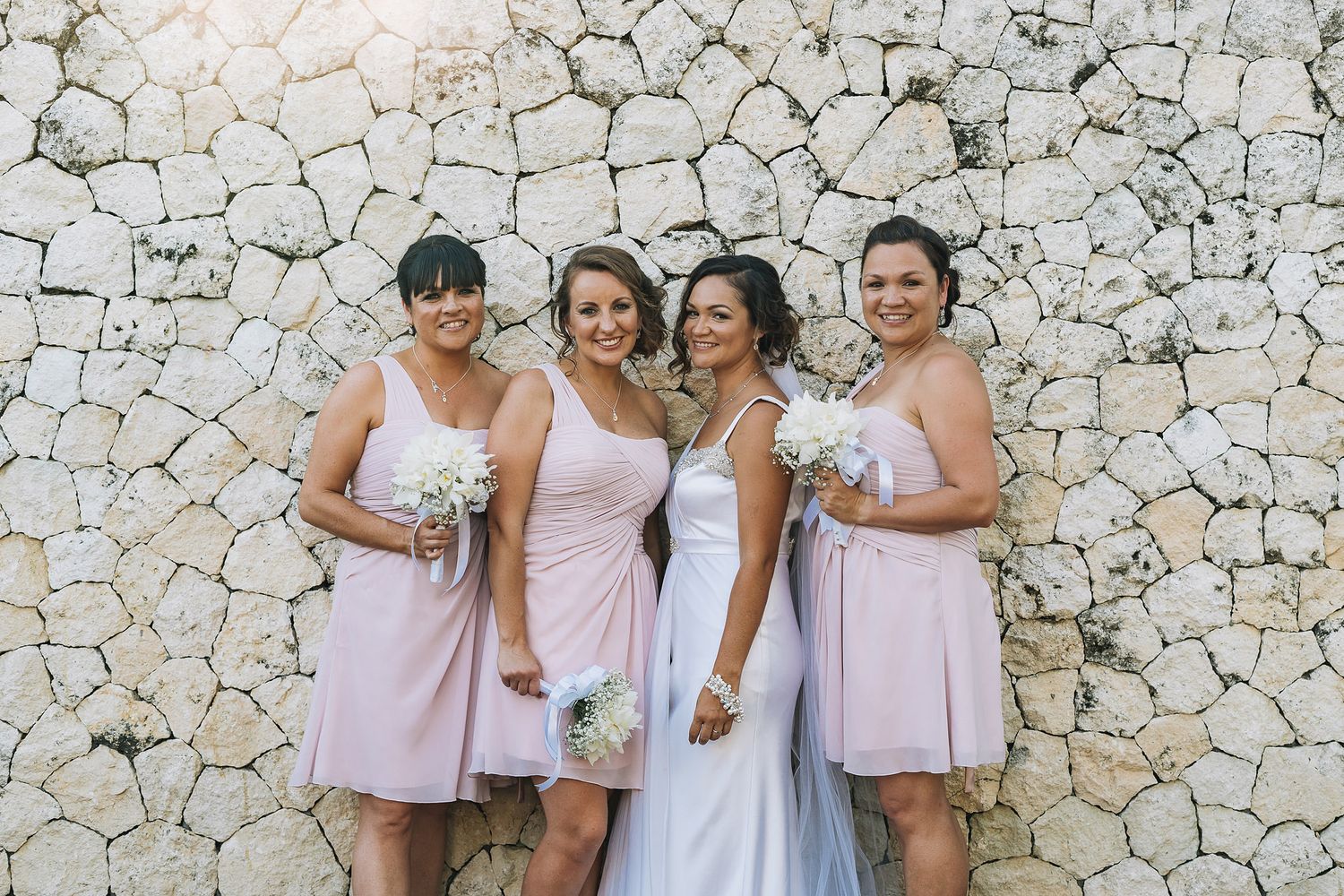 Bridesmaids in pink dresses pose with bride against a stone wall.