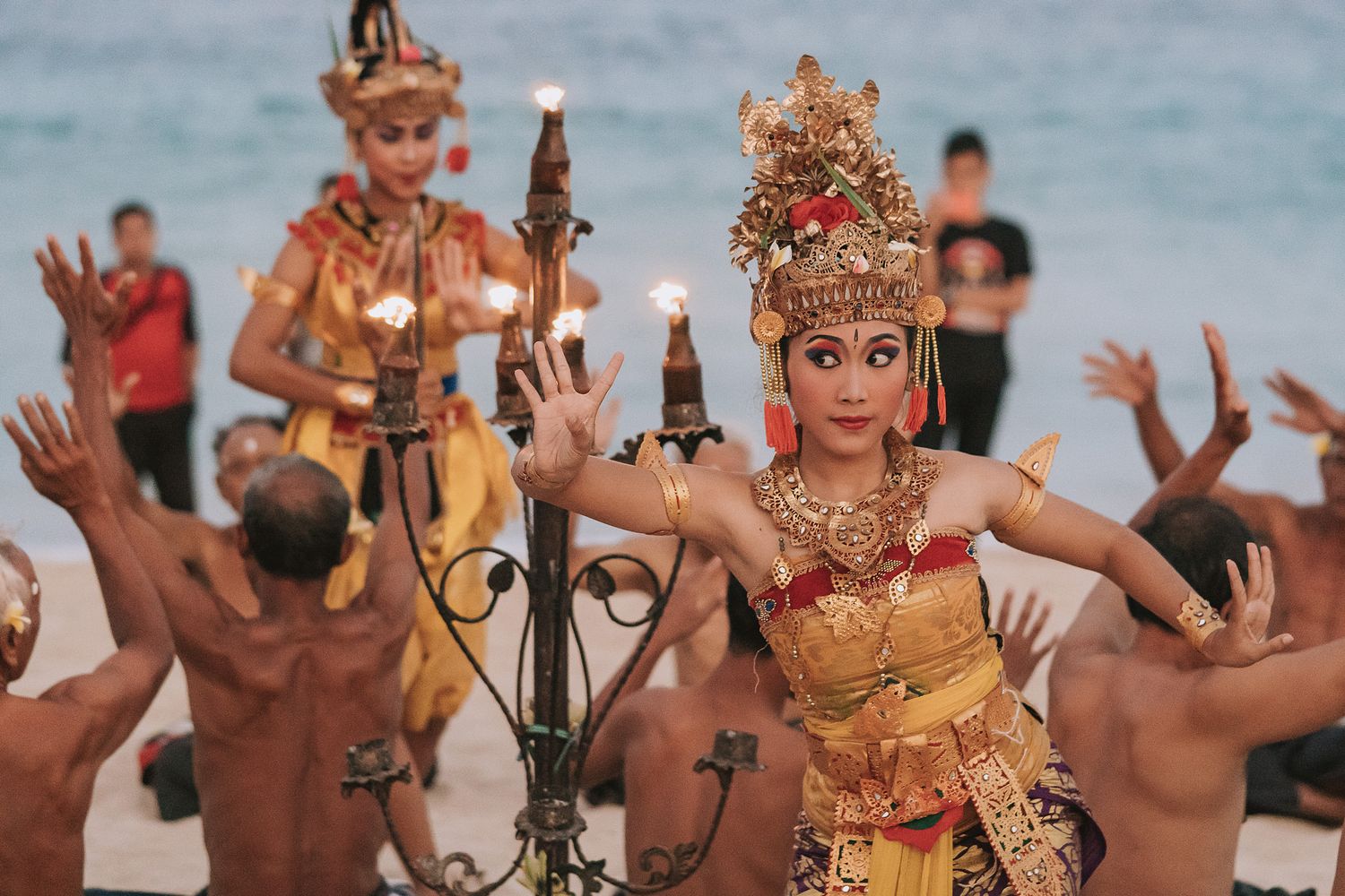 Traditional Balinese dancers perform in ornate costumes and headdresses by the ocean at sunset.