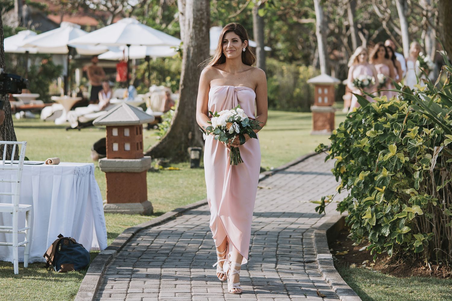 Bridesmaid in pink dress walking down garden path carrying bouquet at outdoor wedding.