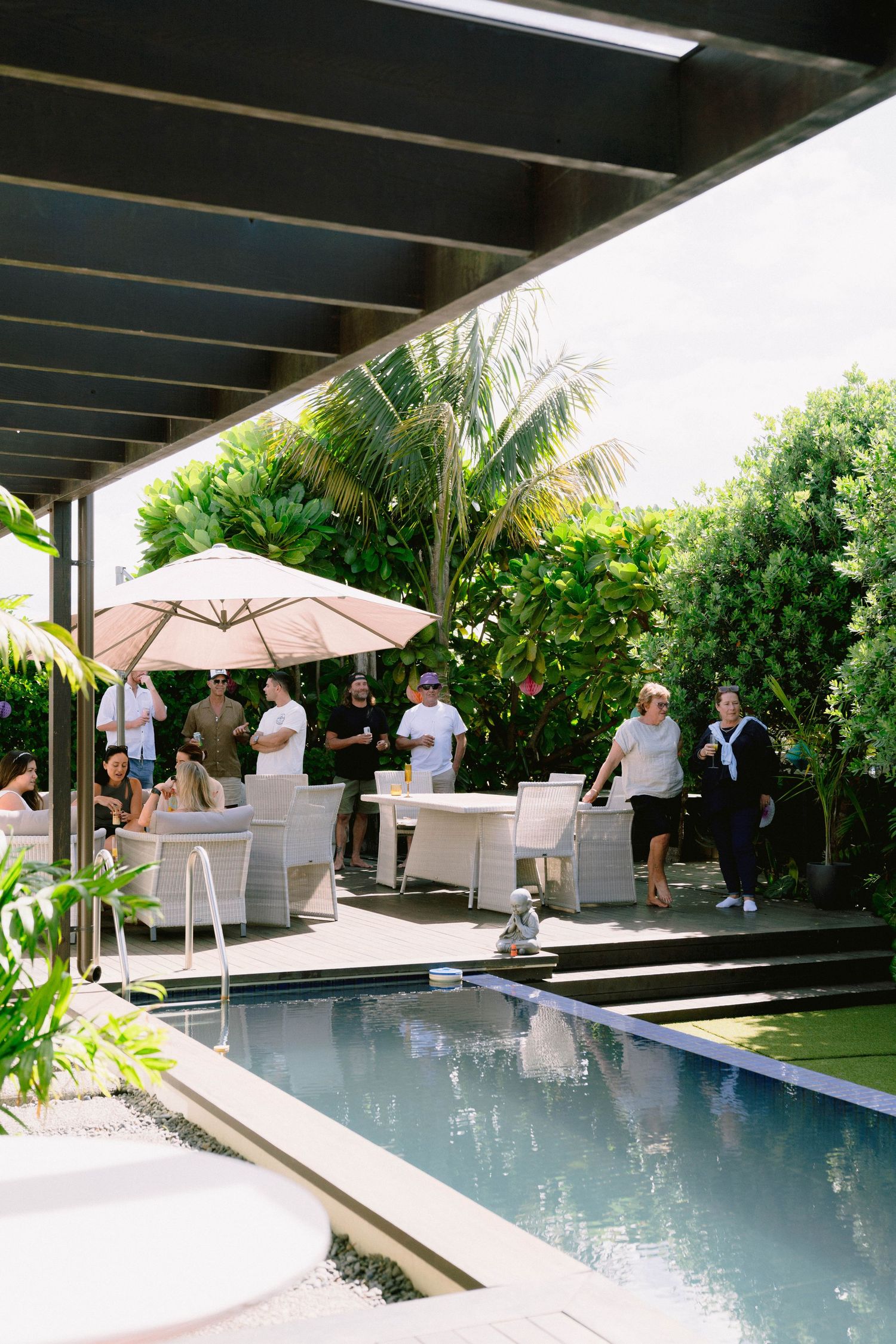People gather around a modern poolside patio with umbrella seating and tropical landscaping in a luxury backyard setting.
