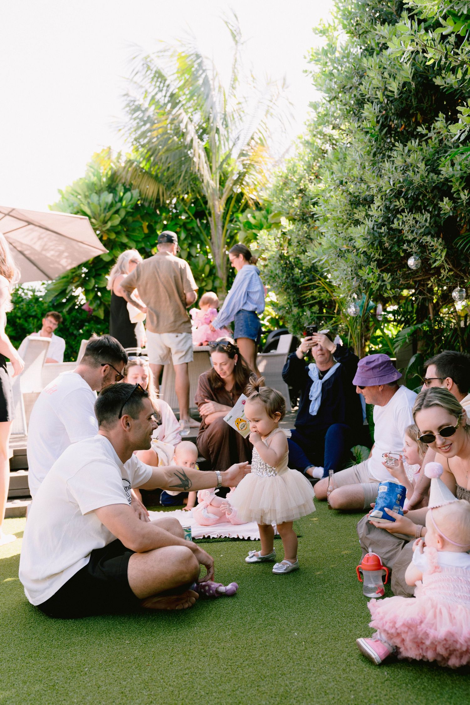 Groups of people gathered outdoors on artificial turf with tropical landscaping at a festive event in pink attire.