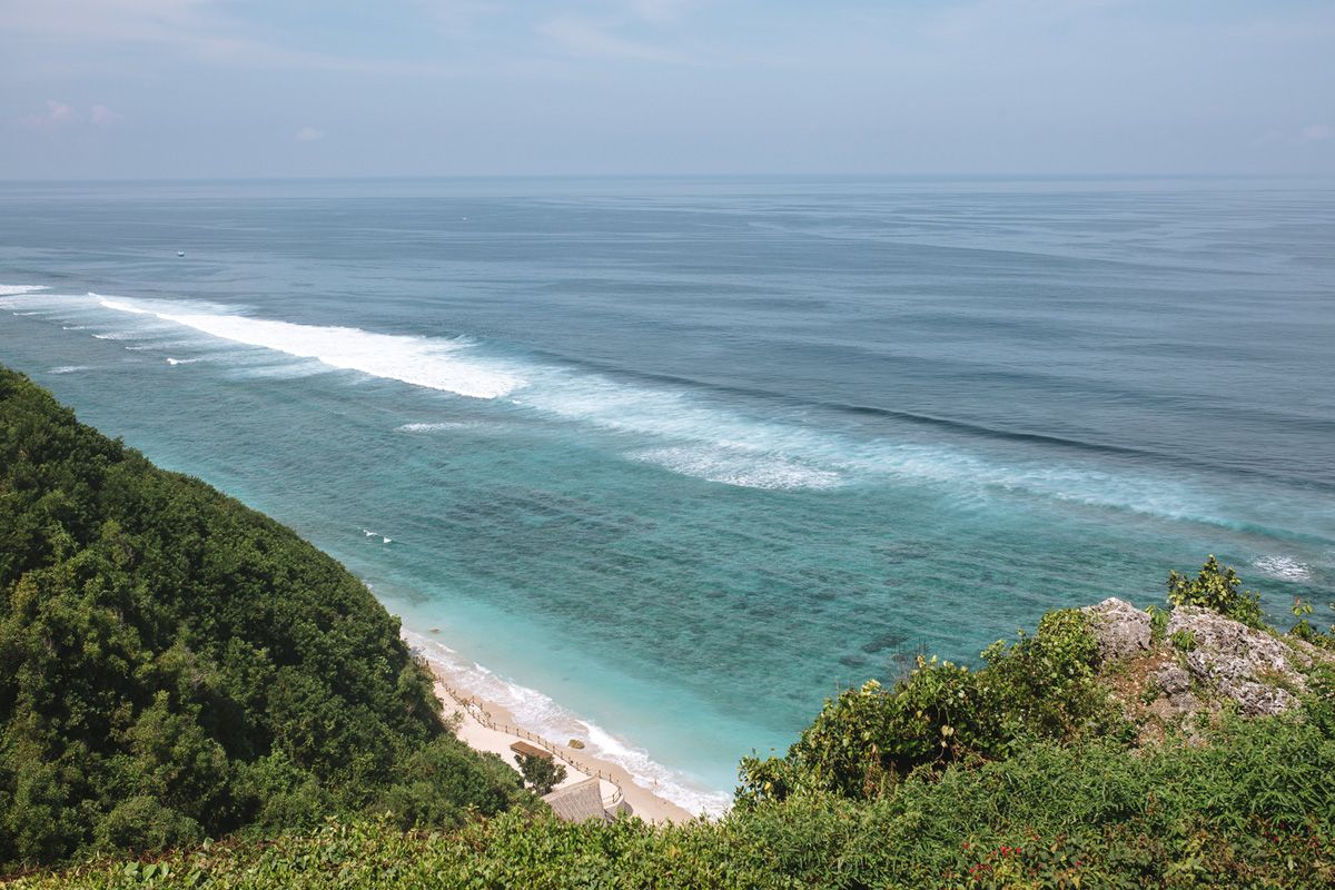 Aerial view of turquoise ocean waves crashing on a tropical beach bordered by lush green cliffs.