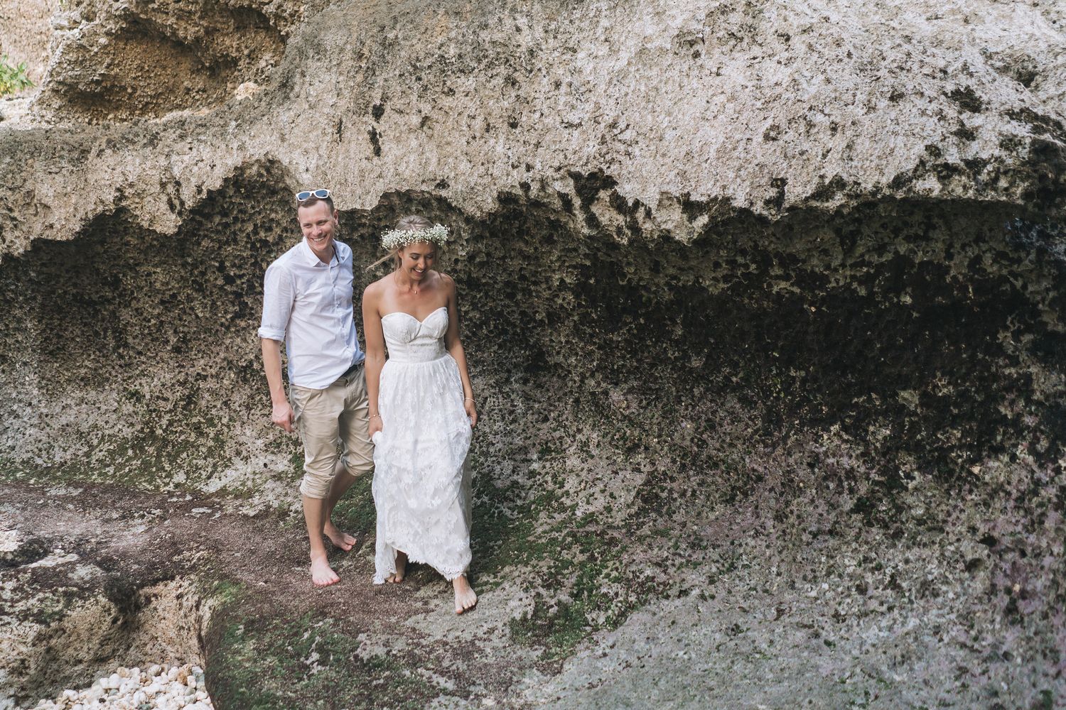A couple stands together on a rocky cliff face wearing wedding attire near the ocean.