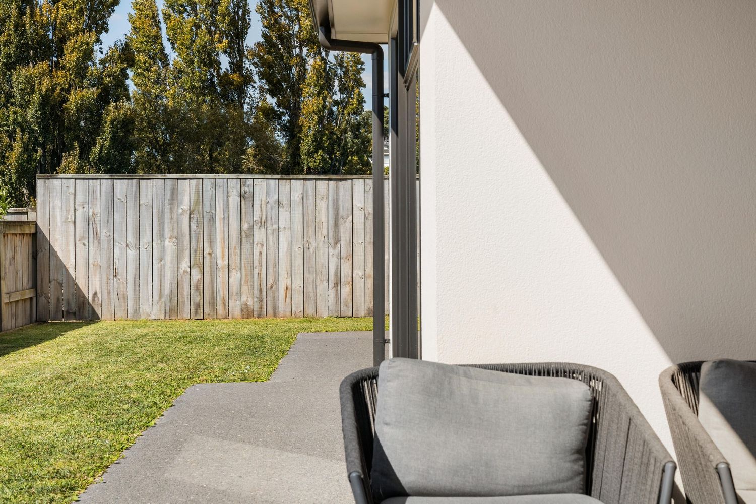 Outdoor patio area with gray cushioned seating against a white wall, wooden fence and green lawn beyond.