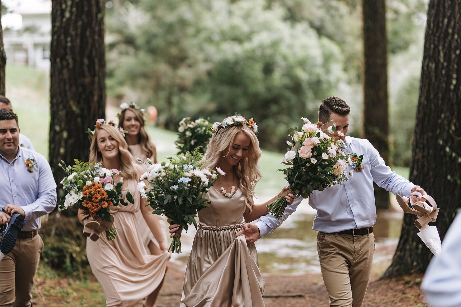 Wedding party in champagne dresses and light blue shirts laugh and dance together in a wooded outdoor setting.