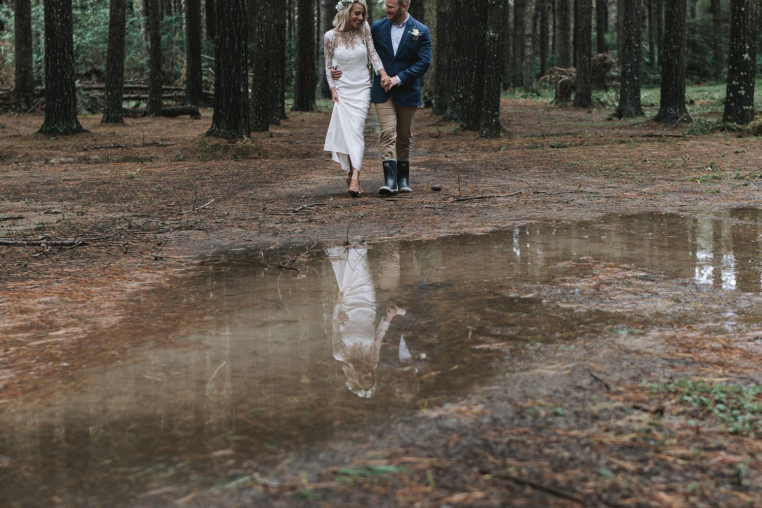 Wedding couple walks together on forest path with their reflection mirrored in puddle on ground.