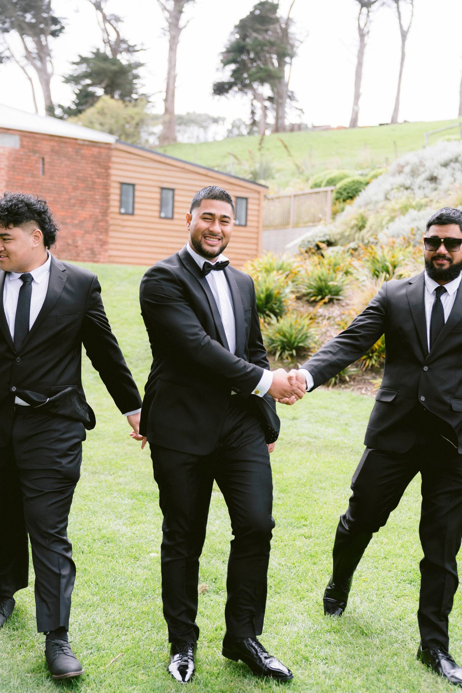 Groomsmen in black suits strike playful poses on a grassy hillside for wedding photos.