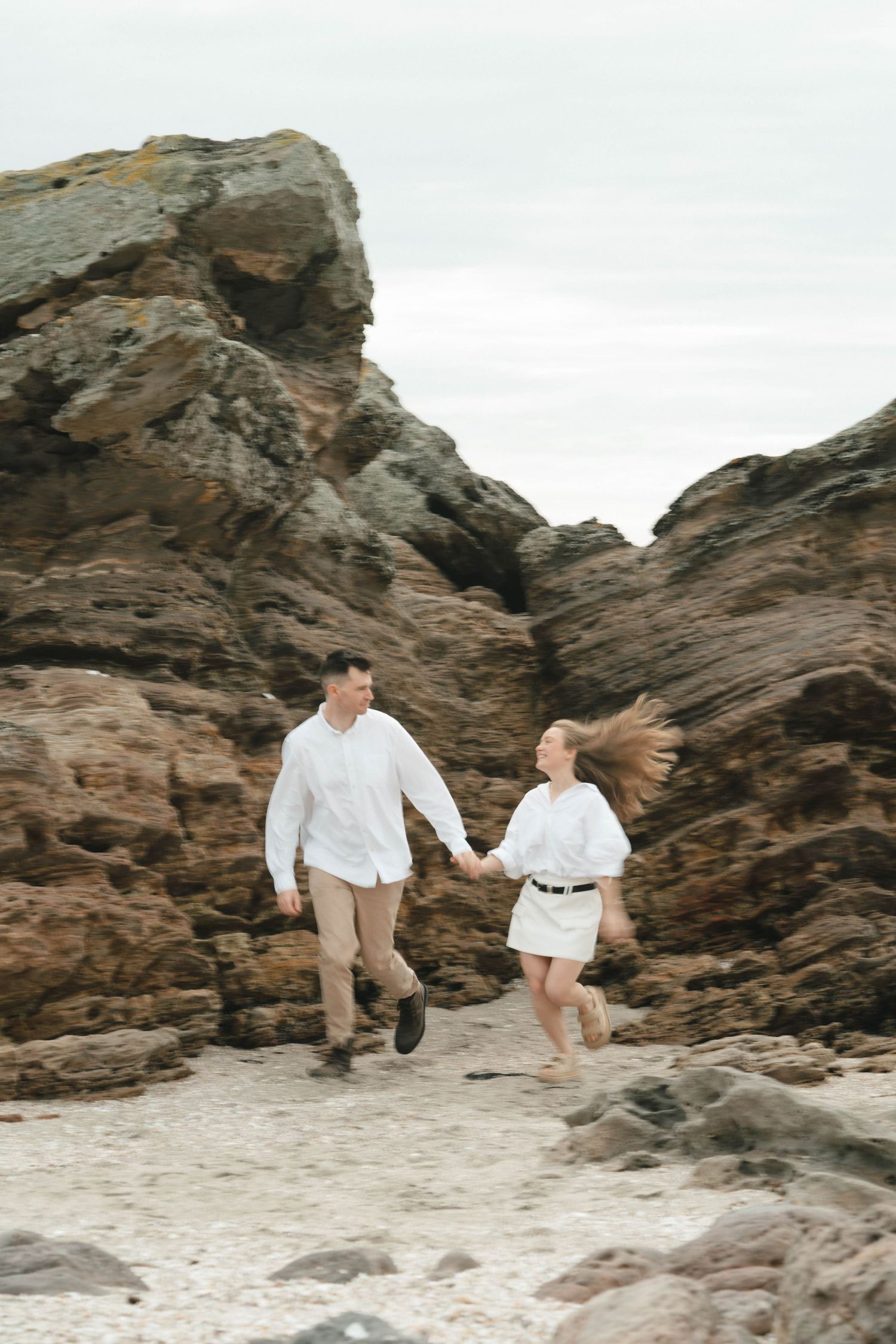 A couple runs playfully together across a rocky beach landscape.