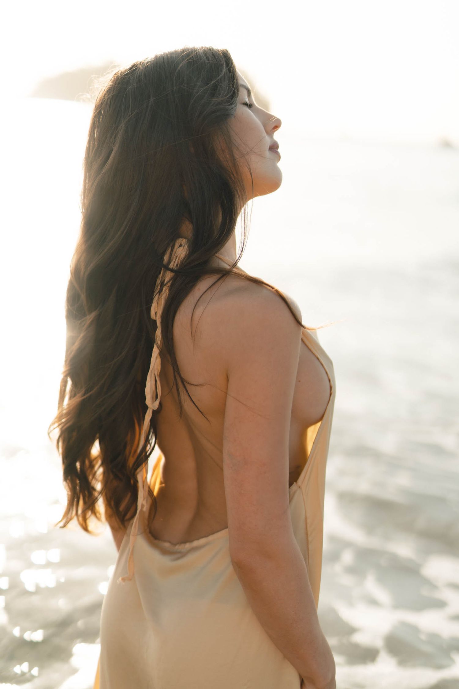 Figure with flowing dark hair poses in cream dress by the ocean at sunset.