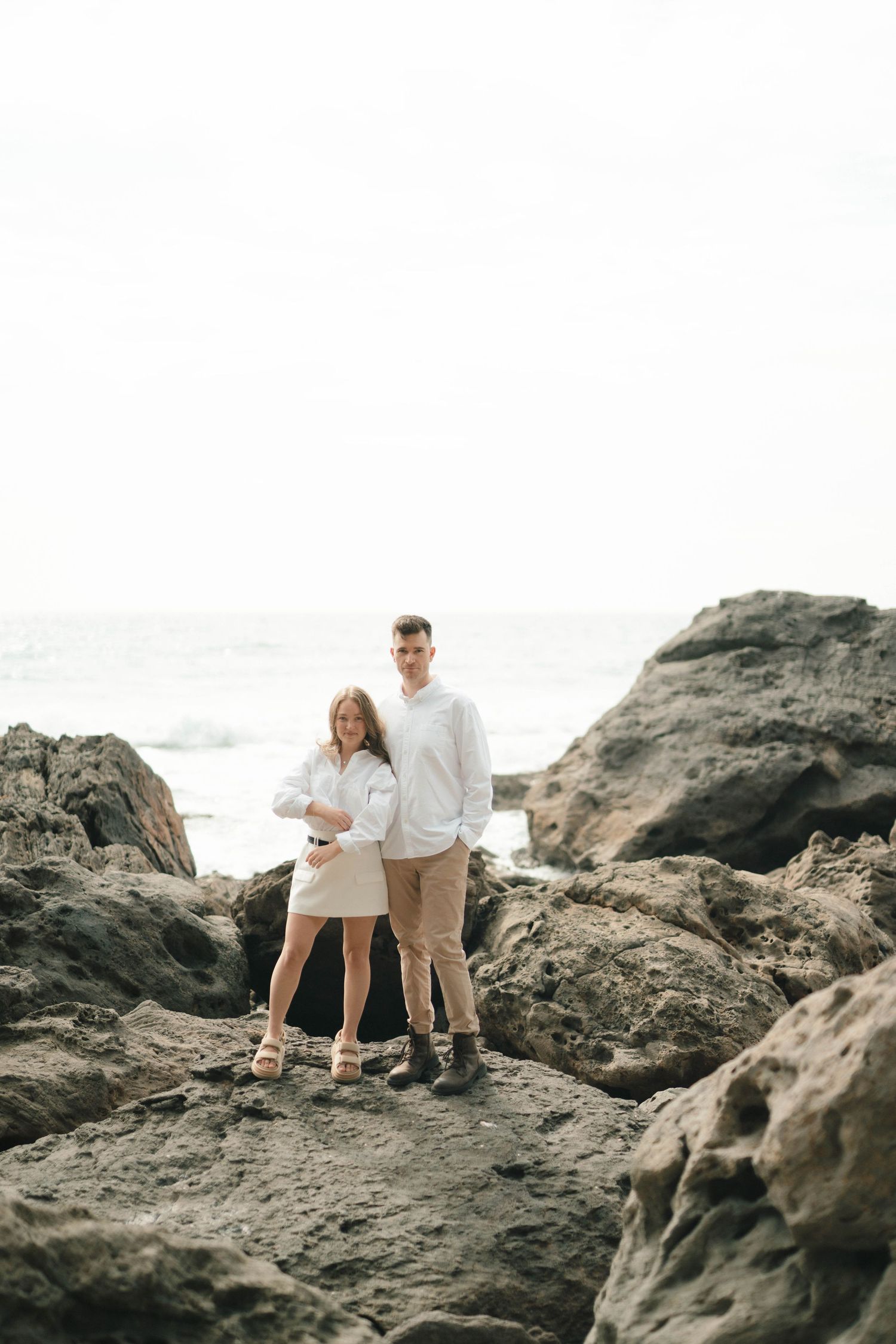 A couple explores rocky coastal cliffs together in white outfits on a bright sunny day.