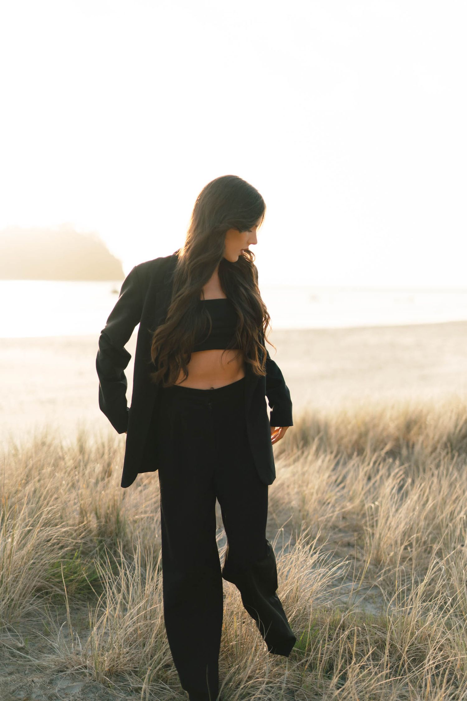 Fashion model in all-black outfit with crop top and blazer poses in golden grass field at sunset.