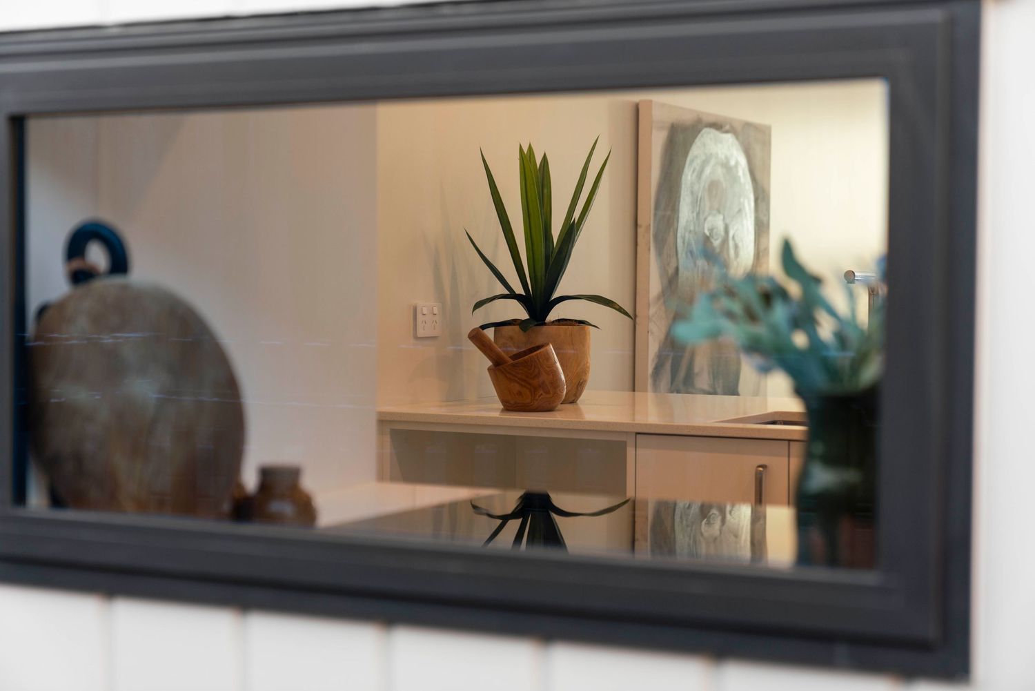 A potted aloe plant and framed artwork reflected in a modern rectangular mirror mounted on a white wall.