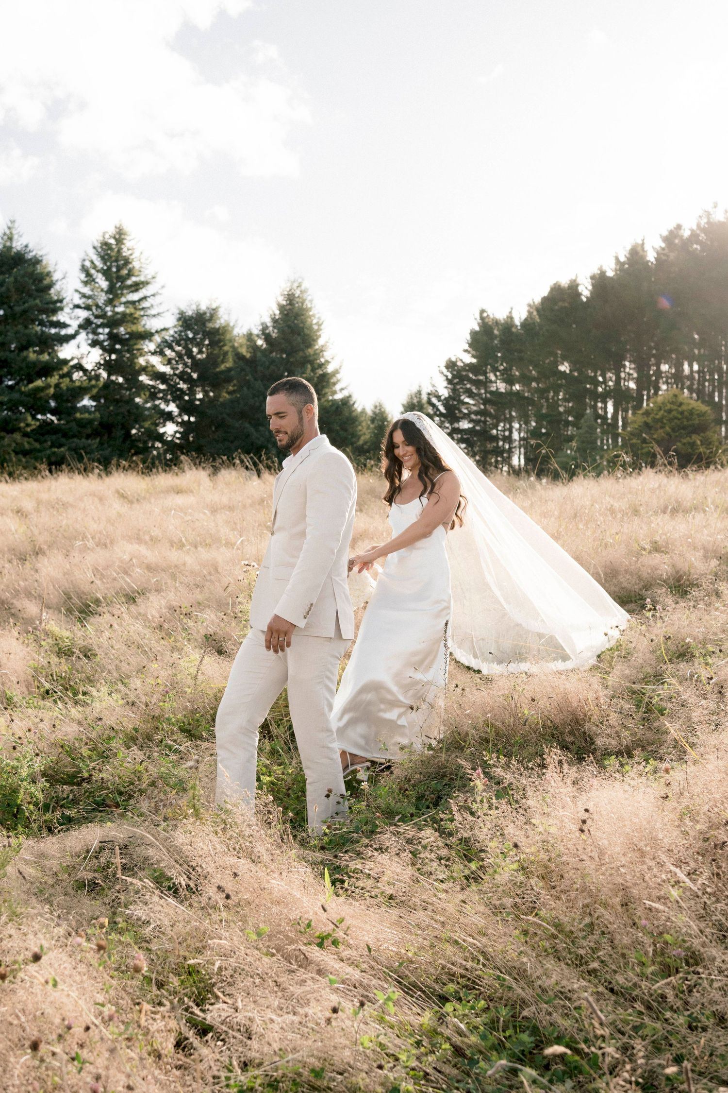 A couple in white outfits walking through a golden field with evergreen trees in background.